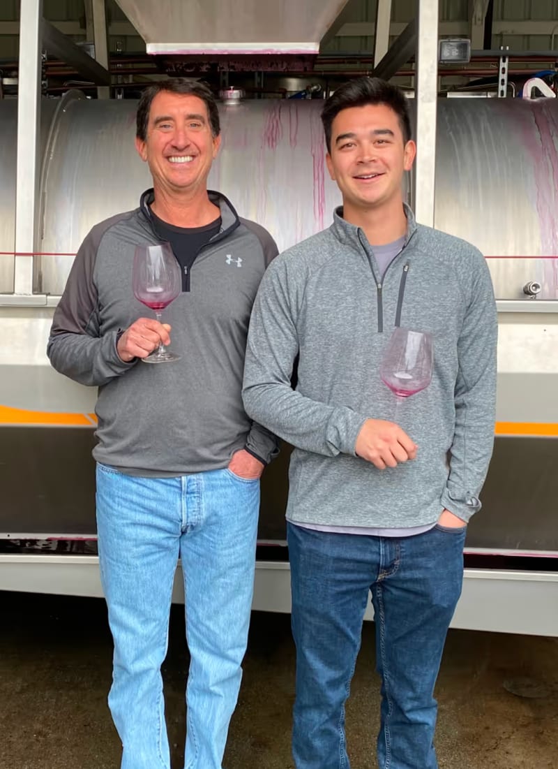 Scott and his dad holding glasses of red wine in front of a stainless steel fermentation tank